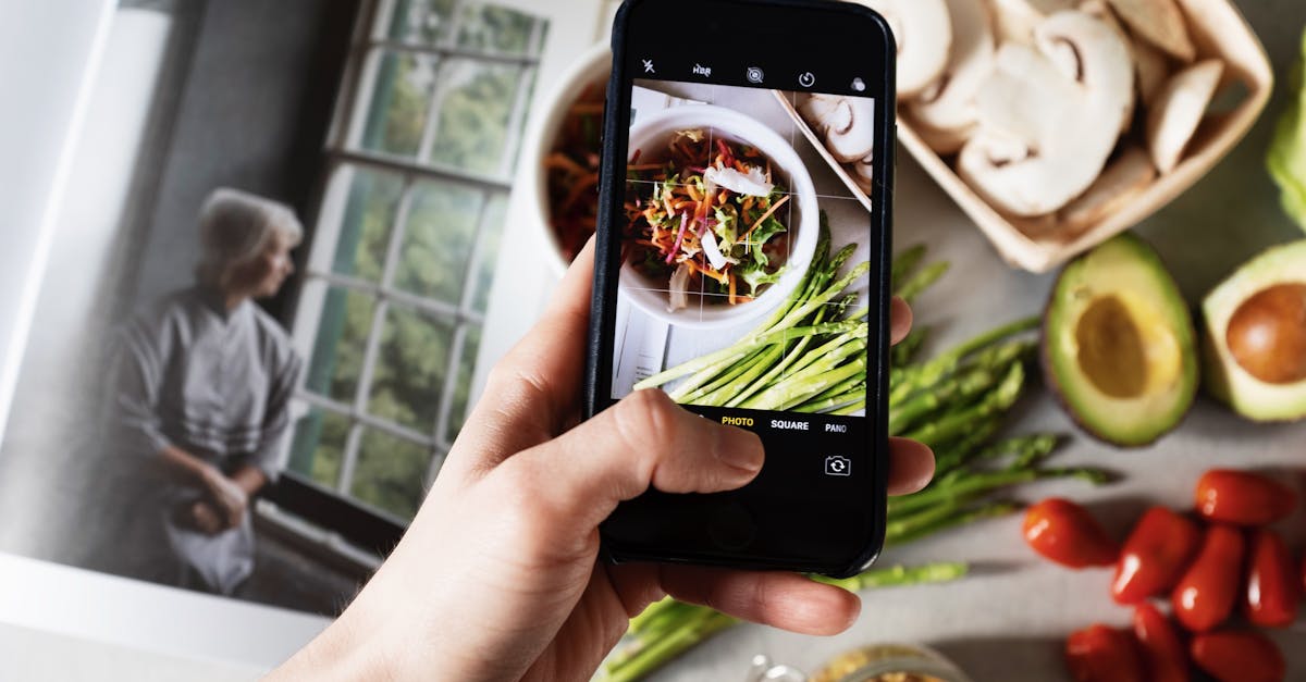 From above view of faceless blogger taking photo on smartphone of fresh tasty salad in white bowl and food placed around on table in modern kitchen