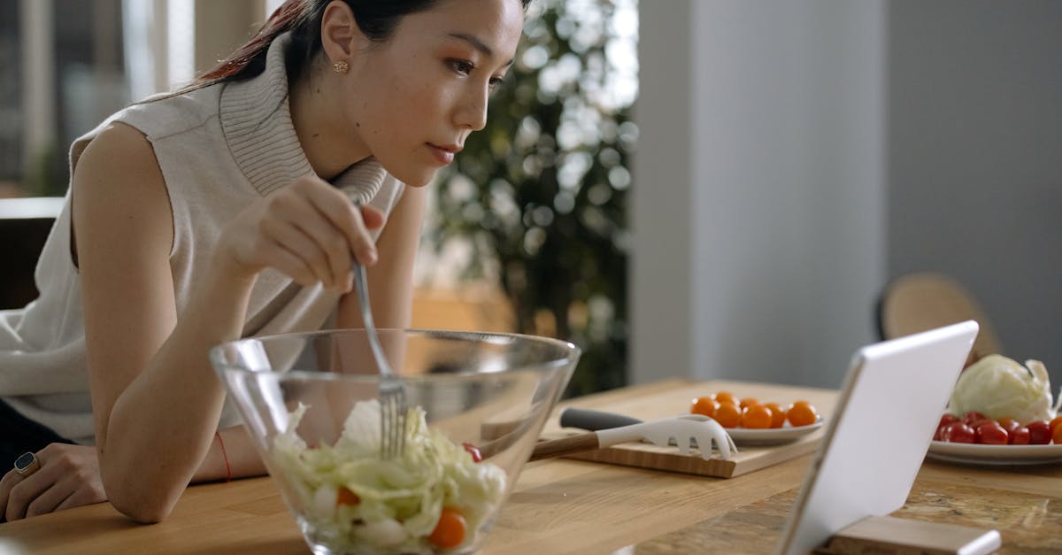 Asian woman preparing a fresh salad while watching a tablet in a cozy kitchen setting.
