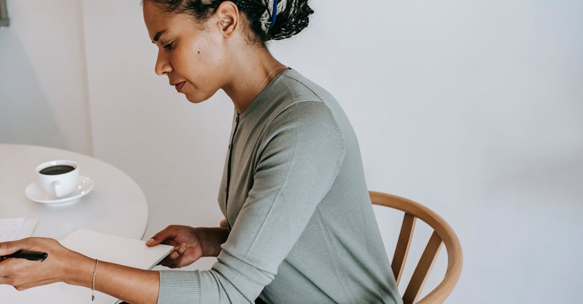Side view crop concentrated ethnic female in formal clothes sitting at table with cup of black coffee and writing notes in clipboard