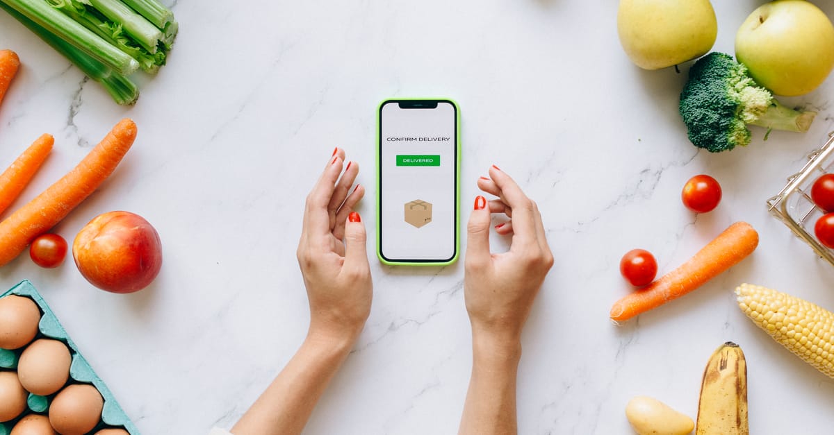 Hands holding smartphone displaying online grocery order amidst fresh vegetables and fruits.