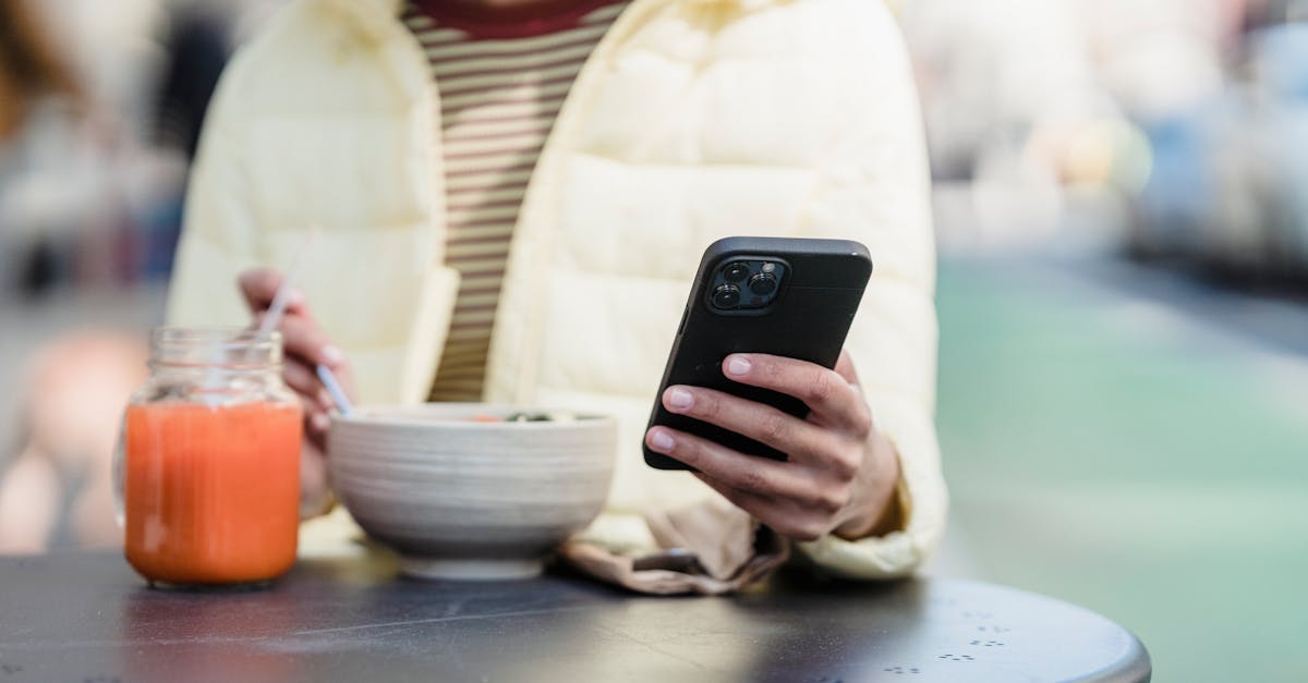 Crop anonymous female surfing internet on cellphone while sitting at urban cafeteria table with food and smoothie in sunlight