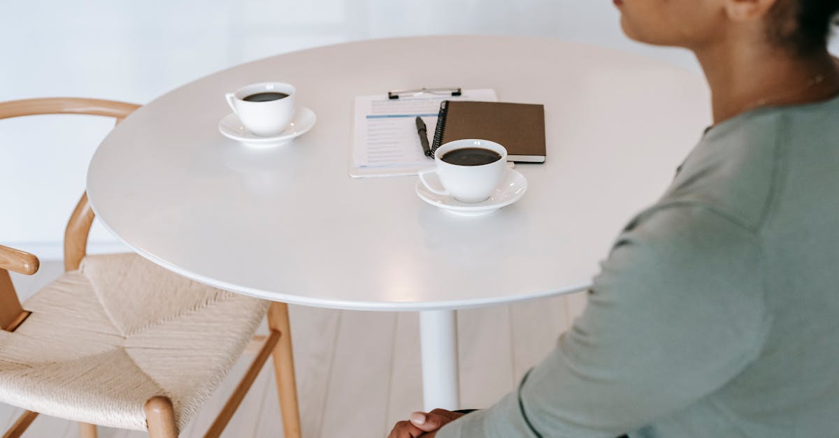 High angle crop ethnic female sitting at round table with cups of aromatic black coffee and diary in light room